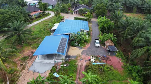 ภาพมุมสูงบ้านหลังคาสีน้ำเงินติดแผงโซล่าเซลล์กลางสวนปาล์ม