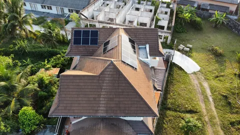 ภาพมุมสูงบ้านหลังคาจั่วสีเข้มที่ติดแผงโซล่าเซลล์ ท่ามกลางแมกไม้