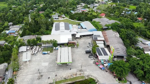ภาพมุมสูงอาคารหลังคาขาวที่ติดตั้งแผงโซล่าเซลล์ท่ามกลางชุมชนและต้นไม้