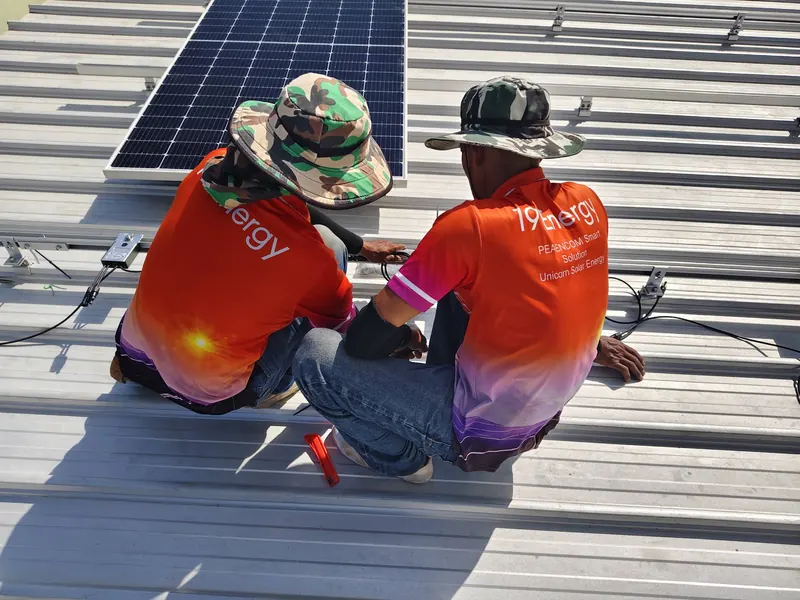 ช่างสองคนสวมเสื้อทีม 19 Energy นั่งต่อสายไฟข้างแผงโซล่าเซลล์บนหลังคาเมทัลชีท ระหว่างติดตั้งรางยึดแผง