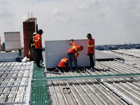 Crew in long-sleeved reflective shirts lifting a solar panel onto the rails of a building roof, with fibreglass walkways between the rows